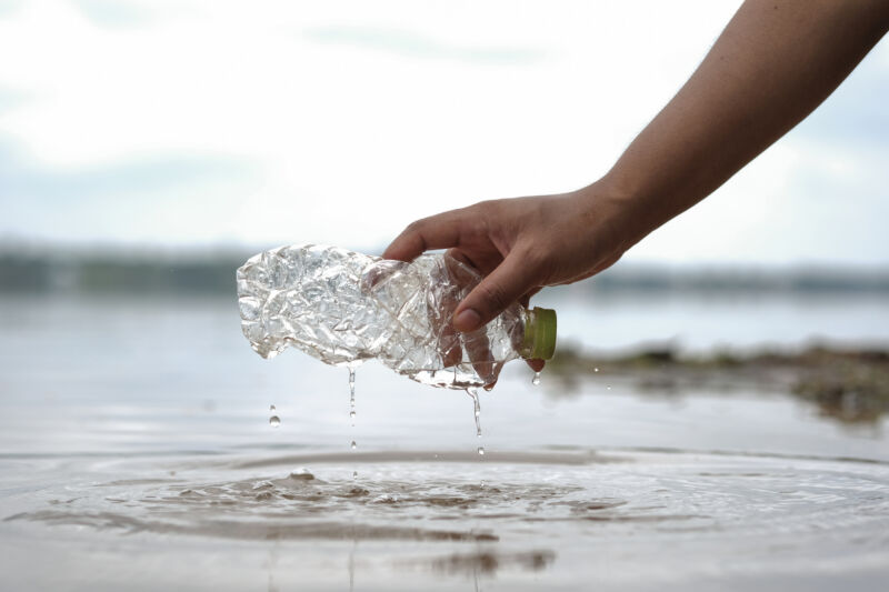 A hand holding a plastic bottle above the sand, with waves in the background.
